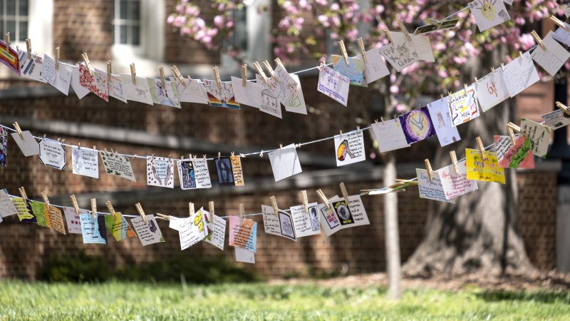 Postcards are strung up on a lawn in front of a tree with pink flowers. After creating and designing postcards with the Carolina staff across campus through the month of March, Arts Everywhere presents all the collected staff postcards in a special exhibit entitled “I was Here: Postcards from the Pandemic,” during the sixth annual Arts Everywhere Day on the campus of the University of North Carolina at Chapel Hill. April 8, 2022. (Jon Gardiner/UNC-Chapel Hill)